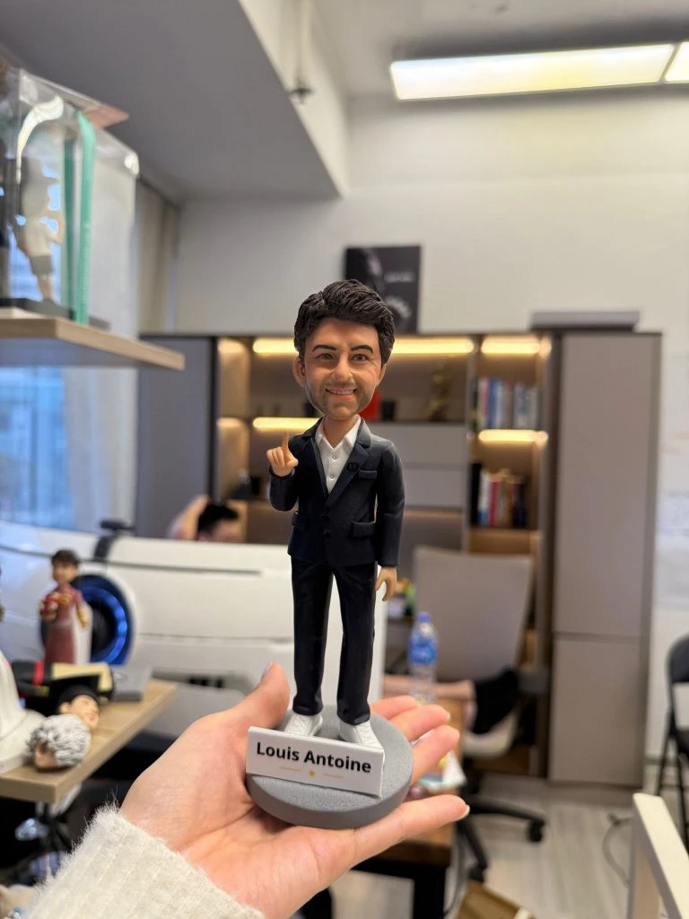 CUTEFIGUREHK 客製化男士公仔。人偶穿著深色西裝外套配白襯衫與白球鞋，展現商務休閒風格。右手舉起食指，面帶自信微笑。灰色圓形底座上帶有「Louis Antoine」專屬名牌及小金星圖案，是完美的升職或職場紀念禮物。