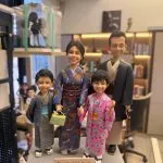 一隻手托著一組客製化的一家四口全家福真人公仔。公仔們穿著傳統日式和服：爸爸穿著深色羽織與淺棕色和服，媽媽穿著華麗的紫色印花和服並提著淺綠色包包，兒子穿著灰色浴衣搭配淺藍色腰帶，女兒穿著粉紅色碎花浴衣。媽媽和女兒牽著手，一家人笑容滿面地站在灰色的長方形底座上，底座前方有一塊寫著「Forever Love」的小牌子。背景為一個帶有層架、書籍與其他公仔的工作室環境。