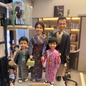 一隻手托著一組客製化的一家四口全家福真人公仔。公仔們穿著傳統日式和服：爸爸穿著深色羽織與淺棕色和服，媽媽穿著華麗的紫色印花和服並提著淺綠色包包，兒子穿著灰色浴衣搭配淺藍色腰帶，女兒穿著粉紅色碎花浴衣。媽媽和女兒牽著手，一家人笑容滿面地站在灰色的長方形底座上，底座前方有一塊寫著「Forever Love」的小牌子。背景為一個帶有層架、書籍與其他公仔的工作室環境。
