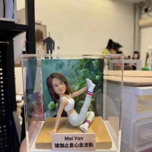 CUTEFIGUREHK 客製化瑜伽主題紀念公仔。一位留著棕色中長髮的女士，身穿白色運動內衣、淺色瑜伽褲及帶有紅白藍條紋的白襪，正在木質底座上優雅地展現單腿伸展的瑜伽姿勢。底座前方設有一塊專屬名牌，印有名字「Mei Yan」及瑜伽哲理金句「瑜伽止息心念波動」。公仔背後配有一幅綠意盎然的自然植物背景圖，整體安放在一個透明的亞加力膠展示盒內。