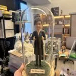 CUTEFIGUREHK 客製化空服員制服公仔。盤髮造型，身穿深色空姐制服配戴金色徽章，做出讚好手勢。公仔放置於帶有仿真草地的木底座玻璃罩內，前方設有印著「ISM Kimmy Chan」的專屬名牌，是送給航空業人士的升職或榮休紀念禮物。