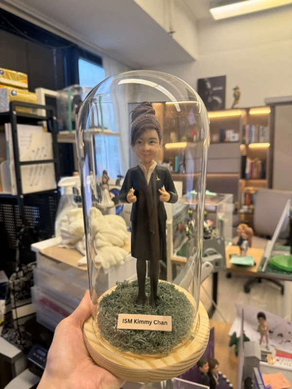 CUTEFIGUREHK 客製化空服員制服公仔。盤髮造型，身穿深色空姐制服配戴金色徽章，做出讚好手勢。公仔放置於帶有仿真草地的木底座玻璃罩內，前方設有印著「ISM Kimmy Chan」的專屬名牌，是送給航空業人士的升職或榮休紀念禮物。