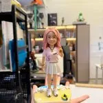 手掌托著的客製公仔：粉紅長髮綁半丸子，穿粉色短袖上衣與白色百褶裙、白色球鞋，配金色手環與項鍊；底座為沙灘與浪花造型，散落貝殼與一只黃藍色救生圈，前方銘牌寫有「跟我一起大啵 今年一定暴富」。背景為室內工作室與層架。