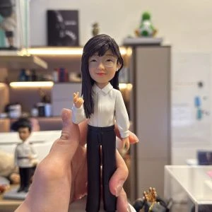手掌托著的客製公仔:長髮戴透明框眼鏡的角色穿白色襯衫與黑色長褲、黑色尖頭鞋,站在圓形白色底座上,右手做比心手勢;背景為室內工作室與層架。