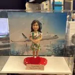 透明展示盒內的客製公仔：穿綠紅花紋洋裝的長髮人物，雙手交疊站在紅色愛心形底座上；前方有小銘牌寫著「Warranty MaMa｜Happy Retirement (2026)」。背板是天空與城市天際線，上方飛過印有「CATHAY PACIFIC」字樣的客機。畫面拍攝於室內辦公環境、可見層架與桌面物品。