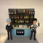 酒吧主題雙人公仔：左側為淺藍髮、西裝與鏈條配件的角色，右側為短黑髮、穿白襯衫與戰術背帶的角色，雙手持道具槍；後方為置滿各色酒瓶的打孔背板層架與小燈飾，中間吧台上有帶蘋果圖示的筆電；吧台前黑色招牌上寫有「永和俠」。