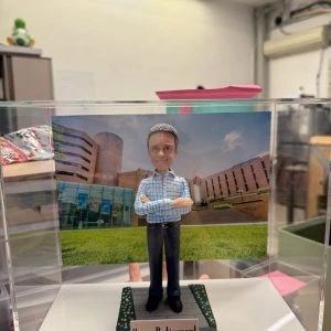 透明展示盒中的客製公仔：短灰髮角色穿淺藍格紋襯衫與深色長褲、黑鞋，雙手交叉站立；腳下為灰色步道與綠植花帶造型底座，前方銘牌寫有「Happy Retirement」。背板是紅磚與玻璃外牆的大樓草坪景色。