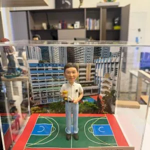 透明展示盒內的畢業主題客製公仔:短髮角色穿白色上衣與淺藍褲、黑鞋,手持金色小獎盃與捲起的證書;腳下是紅綠相間的籃球場樣式底座,上寫字母「C A S C C M C」。右前方銘牌文字為「Lee Ho Lim LOUIS|Happy Graduation 2026 SCC|Love Dad and Mum」。背板為高樓與校舍外觀照片。