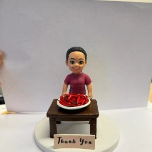 圓形白色底座上的客製公仔：紮馬尾、穿紫紅色上衣的角色站在深木色小桌後方，桌上白盤盛裝滿滿紅色辣椒；前方放置寫有「Thank You」字樣的小牌。背景以白紙遮擋，多為室內環境。