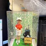 透明展示盒內的客製高爾夫主題公仔：穿白色帽、紅色高爾夫上衣與白裙的人物，手持推桿站在綠色果嶺底座上，腳邊有高爾夫球與球桿；右側放置黑藍配色的球桿袋與球桿組，前方立著小木牌，牌面印有「Christina Our Best Boss」；背板為綠色馬賽克格背景，上方可見「九龍東」與「KED」字樣。場景拍攝於室內辦公環境。
