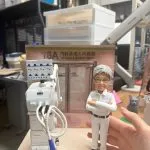 醫護主題客製公仔：短髮、配眼鏡的人物穿全白制服與紅色肩章，雙臂交抱、著黑色工作鞋。左側為帶滾輪的白色醫療設備車，附管線與探頭；背板印有「16A 內科及老人科病房 Medical & Geriatric Ward」。照片拍攝於室內辦公環境，右後方可見小型灰色貓造型擺件。