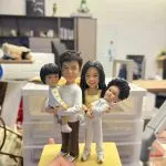 手掌托著的家庭主題客製公仔：一對成人與兩名孩童站在黃色長方形底座上；左側成人抱著短髮孩童，右側成人抱著乳白色連身衣的幼兒。底座前方小銘牌寫有「Happy kong s Family」。背景為室內辦公環境與層架、收納盒。