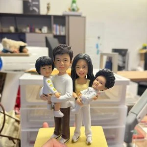 手掌托著的家庭主題客製公仔:一對成人與兩名孩童站在黃色長方形底座上;左側成人抱著短髮孩童,右側成人抱著乳白色連身衣的幼兒。底座前方小銘牌寫有「Happy kong s Family」。背景為室內辦公環境與層架、收納盒。