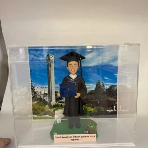 透明展示盒中的畢業主題公仔:角色穿黑色學士服與方帽,手抱藍色學位夾;腳下綠色草地造型底座,旁有書本與布袋裝飾。背板為藍天空下的校園景色與鐘樓。前方銘牌寫有「The University of British Columbia 2026|Alton Ho」。