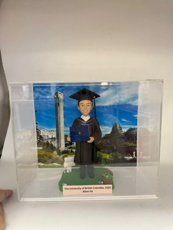 透明展示盒中的畢業主題公仔：角色穿黑色學士服與方帽，手抱藍色學位夾；腳下綠色草地造型底座，旁有書本與布袋裝飾。背板為藍天空下的校園景色與鐘樓。前方銘牌寫有「The University of British Columbia 2026｜Alton Ho」。