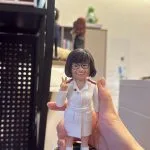 手掌托著的護理主題客製公仔：短髮戴眼鏡的人物穿全白制服，紅色肩章，右手比出勝利手勢、面帶微笑；腳踩白色球鞋，站在深綠圓形底座上，邊緣點綴粉黃小圓點。背景為室內辦公環境與層架。