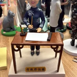 客製公仔站在中式木桌後方:短髮、戴眼鏡、微笑的角色穿海藍色花卉圖案上衣與深色長褲、高跟鞋,右手持毛筆,桌上放白紙、毛筆筒與墨條;底座前方金色小牌寫著「母親節快樂」。背景有其他公仔與擺設,場景為室內工作臺面。