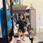 手掌托著的籃球主題客製公仔：短髮人物穿黑色POLO上衣與黑色短褲、白色運動鞋，雙手叉腰，右腳踩在橙藍配色的Wilson 3x3籃球上；左手戴藍色智能手錶。底座為灰色蜂巢紋石板；背景是室內辦公環境與層架、文件夾與書櫃。