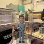透明展示盒中的客製公仔：短髮角色穿藍灰色警務制服與黑皮帶、黑鞋，右手比讚站立；胸前有「POLICE」與階級肩章；石紋底座前粉色銘牌印有祝福語。背景為室內工作室。
