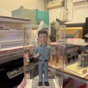 透明展示盒中的客製公仔:短髮角色穿藍灰色警務制服與黑皮帶、黑鞋,右手比讚站立;胸前有「POLICE」與階級肩章;石紋底座前粉色銘牌印有祝福語。背景為室內工作室。