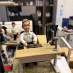 手掌托著的客製公仔：短灰髮、微笑的角色穿白色襯衫坐在辦公椅上，面前是木色辦公桌，擺有黑色鍵盤與滑鼠、側邊裝有銀灰色支架的黑色螢幕；公仔與桌面置於仿木紋底座上。背景為室內辦公空間與層架。