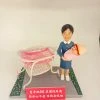 場景公仔置於灰色石板紋底座：穿藍色制服與白色鞋的角色懷抱粉色包裹的新生兒，旁邊有白粉色新生兒推床與透明床罩；底座兩側以綠色花帶點綴，前方紅色銘牌以白字寫上中文祝福語（此處不逐字轉錄），背景為淺色牆面。