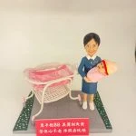場景公仔置於灰色石板紋底座：穿藍色制服與白色鞋的角色懷抱粉色包裹的新生兒，旁邊有白粉色新生兒推床與透明床罩；底座兩側以綠色花帶點綴，前方紅色銘牌以白字寫上中文祝福語（此處不逐字轉錄），背景為淺色牆面。