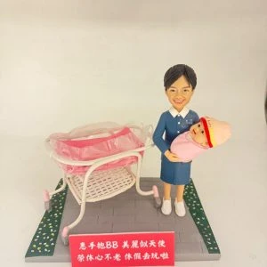 場景公仔置於灰色石板紋底座：穿藍色制服與白色鞋的角色懷抱粉色包裹的新生兒，旁邊有白粉色新生兒推床與透明床罩；底座兩側以綠色花帶點綴，前方紅色銘牌以白字寫上中文祝福語（此處不逐字轉錄），背景為淺色牆面。