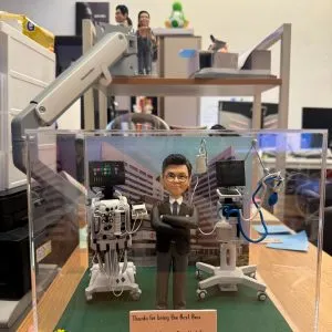 透明展示盒中的場景公仔:角色穿深色西裝、雙臂交叉站立;左右兩側各有一台附螢幕與多條管線的醫療設備推車;底板為草綠色並點綴小花片,前方銅色銘牌寫有「Thanks for being the Best Boss / Paediatric Intensive Care Unit Team / Department of Paediatrics, PWH / Year 2026」。背板為醫院外觀影像