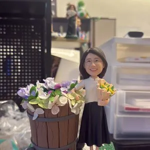 手持展示的一尊場景公仔:角色穿淺色上衣與深色長裙,手臂抱著小花束;身旁是一只帶輪的木桶,桶內盛滿紫、粉、白與綠色的繡球花;腳下為灰色石板路造型底座,兩側綠地點綴小花,前方白色銘牌以黑字寫有「幸福快樂」。