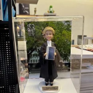 透明展示盒中的場景公仔：角色穿黑色法袍、搭配金色捲曲律師假髮與白色領巾，雙手持藍色封面的法典手冊（封面可見「Prosecution Code」字樣）；腳踩白色圓形底座，前方金色銘牌寫有「Maggie Yang / Happy retirement」。背板為綠樹與建築景色，左上角有黑白書法字樣。