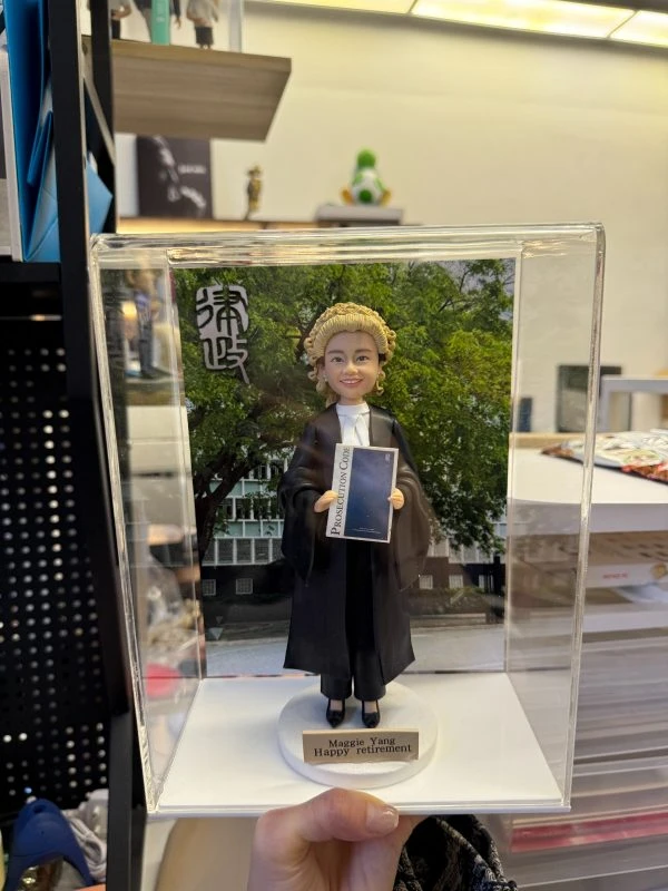 透明展示盒中的場景公仔：角色穿黑色法袍、搭配金色捲曲律師假髮與白色領巾，雙手持藍色封面的法典手冊（封面可見「Prosecution Code」字樣）；腳踩白色圓形底座，前方金色銘牌寫有「Maggie Yang / Happy retirement」。背板為綠樹與建築景色，左上角有黑白書法字樣。