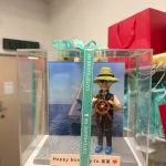 透明展示盒中的場景公仔：角色頭戴草帽與墨鏡，穿深色背心與藍色鞋，雙手握著棕色船舵；背板為藍色海面與白色帆船圖像；底座為灰色石板紋，前方長條銘牌寫有「Happy birthday to 弟弟」