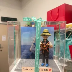 透明展示盒中的場景公仔:角色頭戴草帽與墨鏡,穿深色背心與藍色鞋,雙手握著棕色船舵;背板為藍色海面與白色帆船圖像;底座為灰色石板紋,前方長條銘牌寫有「Happy birthday to 弟弟」