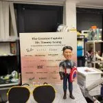 場景公仔置於灰色石板紋大底座上，前方有一副黃色外框的大型飛行員墨鏡模型；右側角色著藍白紅配色的隊長風服裝並持圓形盾牌。背板印有「The Greatest Captain – Mr. Tommy Leung」「Congratulations on a well‑deserved retirement!」等文字與多個精品品牌字樣；底部另有小銘牌