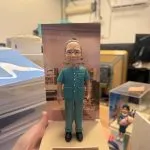 手掌托起的場景公仔：角色穿藍綠色短袖醫護制服與同色長褲、配戴眼鏡與黑色鞋，髮髻紮起；站在米色砂感長方形底座上，背板為室內科室或藥局場景。前方金色銘牌印有多行中文：「全哥！榮休之喜 多謝勞苦功高 律敦治及鄧肇堅醫院藥劑部」。