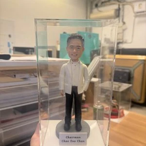 透明展示盒中的場景公仔:角色著白色西裝外套與白襯衫、搭配黑長褲與黑鞋;站在灰色圓形底座上,前方白色銘牌印有「Chairman / Chae Soo Chan」文字。背景為室內工作室與收納設備。