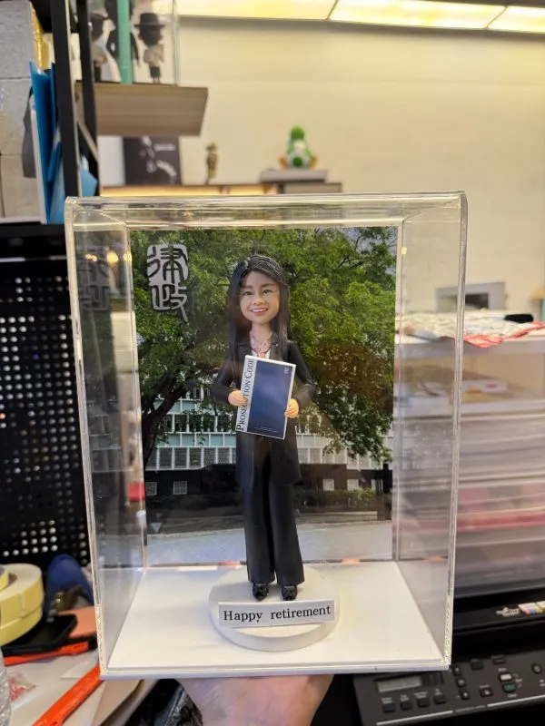 透明展示盒中的場景公仔：身著黑色西裝與黑鞋的角色手持深藍色文件手冊，站在圓形灰色底座上；前方白色小銘牌寫有「Happy retirement」；背板為綠樹與現代建築外牆，左上角有直排漢字。