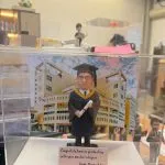 透明展示盒中的場景公仔：角色穿黑色學士袍與學位帽、披金藍配色的領巾，手持綁紅繩的卷軸；背板為現代感校園建築與藍天雲彩；前方米色銘牌以英文寫有畢業祝福與署名（此處不逐字轉錄）。