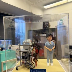 透明展示盒中的場景公仔：角色穿淺藍色醫護制服、配白鞋與識別證，雙手於身前；旁邊是一台帶有監視器與多條電線的灰色醫療設備推車；底板為米黃色質感，前方藍色銘牌寫有「To our dearest JoJo / PYNEH ICU 2026」等字樣。背景為室內辦公與收納區。