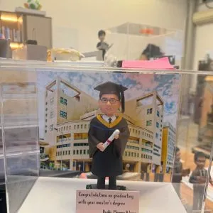 透明展示盒中的場景公仔:角色穿黑色學士袍與學位帽、披金藍配色的領巾,手持綁紅繩的卷軸;背板為現代感校園建築與藍天雲彩;前方米色銘牌以英文寫有畢業祝福與署名(此處不逐字轉錄)。