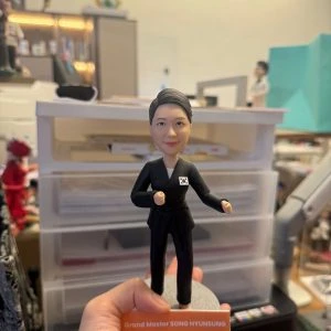 手持展示的一尊武術主題公仔：角色穿黑色道服與黑帶，胸前貼有小型國旗貼章，雙手呈出拳姿，腳下為灰色圓形底座；前方橘色銘牌印有英文字職稱與姓名（此處不逐字轉錄），背景為工作桌與收納抽屜。