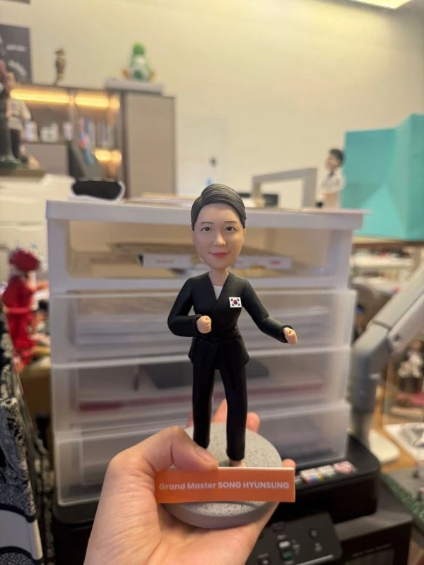 手持展示的一尊武術主題公仔：角色穿黑色道服與黑帶，胸前貼有小型國旗貼章，雙手呈出拳姿，腳下為灰色圓形底座；前方橘色銘牌印有英文字職稱與姓名（此處不逐字轉錄），背景為工作桌與收納抽屜。