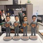 單一公仔特寫：穿灰色西裝與黑領帶的角色站在灰色圓形底座上，左手持寫有「BEAM」字樣的手牌，右手比讚；前方金屬色銘牌以英文寫有稱呼與「Happy Retirement」（此處不逐字轉錄），背景是辦公室與書架。
