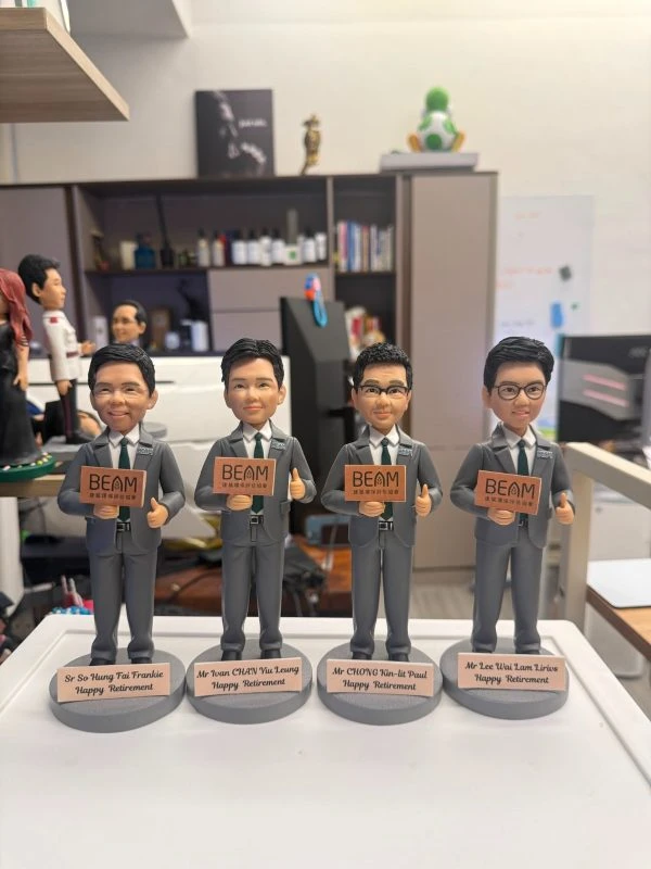 單一公仔特寫：穿灰色西裝與黑領帶的角色站在灰色圓形底座上，左手持寫有「BEAM」字樣的手牌，右手比讚；前方金屬色銘牌以英文寫有稱呼與「Happy Retirement」（此處不逐字轉錄），背景是辦公室與書架。