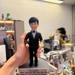 手持展示的一尊指揮主題公仔:角色穿黑色西裝與黑色領結,雙手持細長指揮棒,站在灰色圓形底座上;前方白色銘牌以英文寫有「Best Music Conductor」「Class of 2026」等字樣,並列出多名中文姓名(此處不逐字轉錄);背景為辦公室與書架。