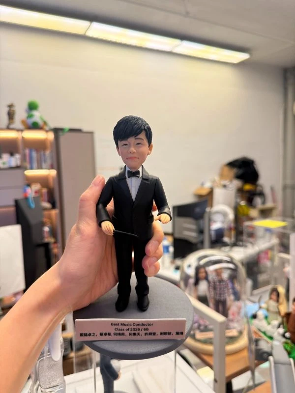 手持展示的一尊指揮主題公仔：角色穿黑色西裝與黑色領結，雙手持細長指揮棒，站在灰色圓形底座上；前方白色銘牌以英文寫有「Best Music Conductor」「Class of 2026」等字樣，並列出多名中文姓名（此處不逐字轉錄）；背景為辦公室與書架。