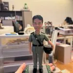 手持展示的一尊制服主題公仔：角色穿淺綠色制服與黑色皮帶，肩章細節可見，右手持深色軍帽站在灰色石板紋底座上；底座邊緣有綠色與黃色小花裝飾，前方白色銘牌以中文寫有職稱與祝福語（不逐字轉錄）；背景為辦公室與收納抽屜。