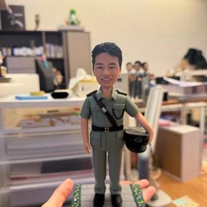 手持展示的一尊制服主題公仔:角色穿淺綠色制服與黑色皮帶,肩章細節可見,右手持深色軍帽站在灰色石板紋底座上;底座邊緣有綠色與黃色小花裝飾,前方白色銘牌以中文寫有職稱與祝福語(不逐字轉錄);背景為辦公室與收納抽屜。