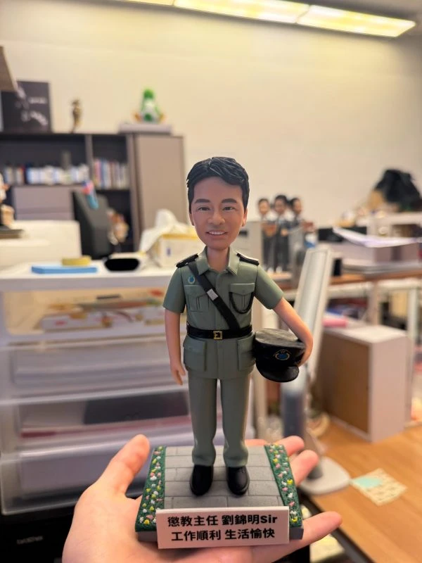 手持展示的一尊制服主題公仔：角色穿淺綠色制服與黑色皮帶，肩章細節可見，右手持深色軍帽站在灰色石板紋底座上；底座邊緣有綠色與黃色小花裝飾，前方白色銘牌以中文寫有職稱與祝福語（不逐字轉錄）；背景為辦公室與收納抽屜。